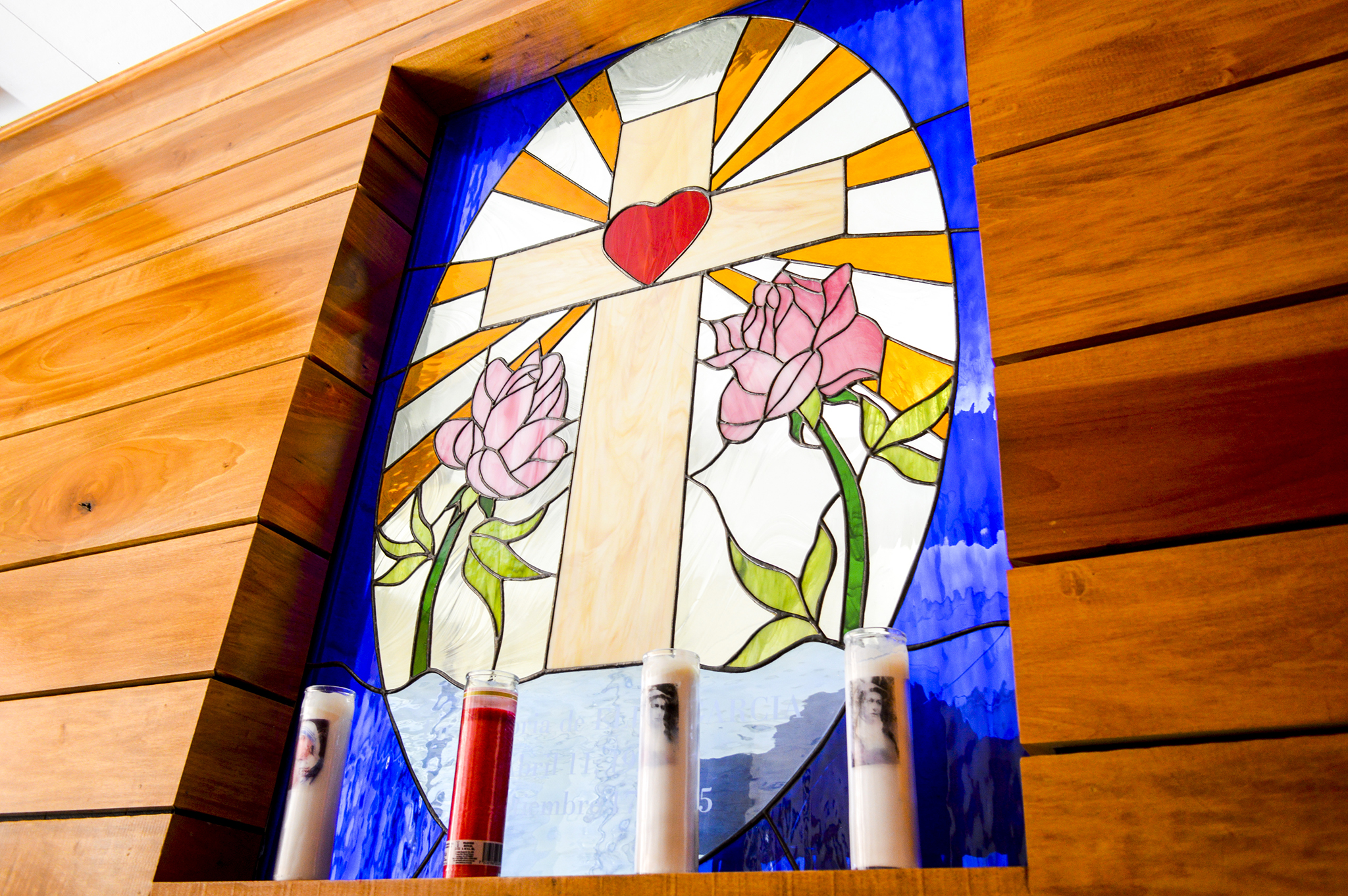 A wood-paneled wall harbors a stained glass mosaic window of a cross. In the middle of the cross is a red heart and two roses are on either side of the cross. Light and dark blue frame the outside of the cross. Candles line the bottom of the cross.