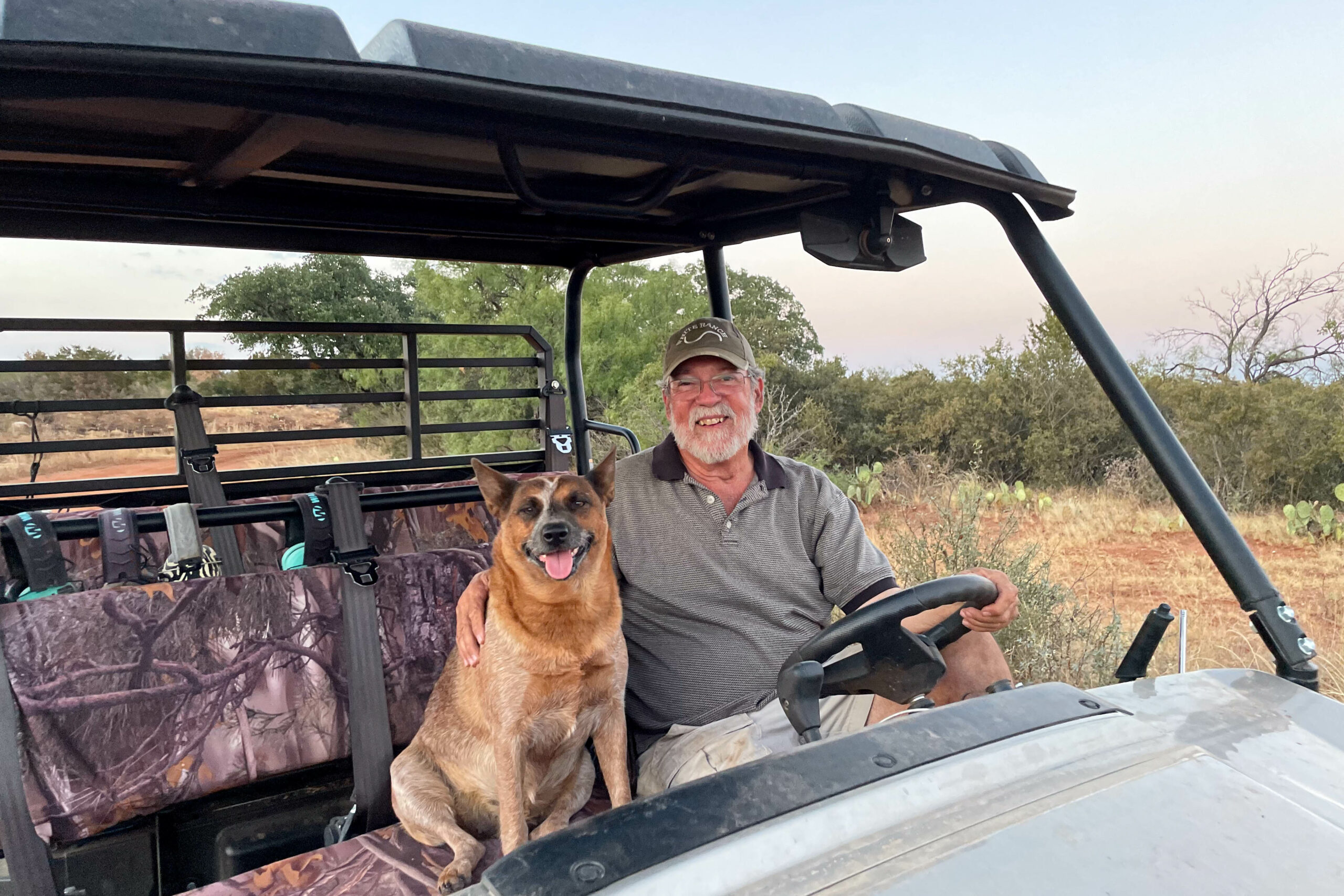 Medium shot of Brian Wright and his dog sitting in an open-air ranch vehicle. Brian Wright wears a White Ranch baseball cap and has his arm on his dog.