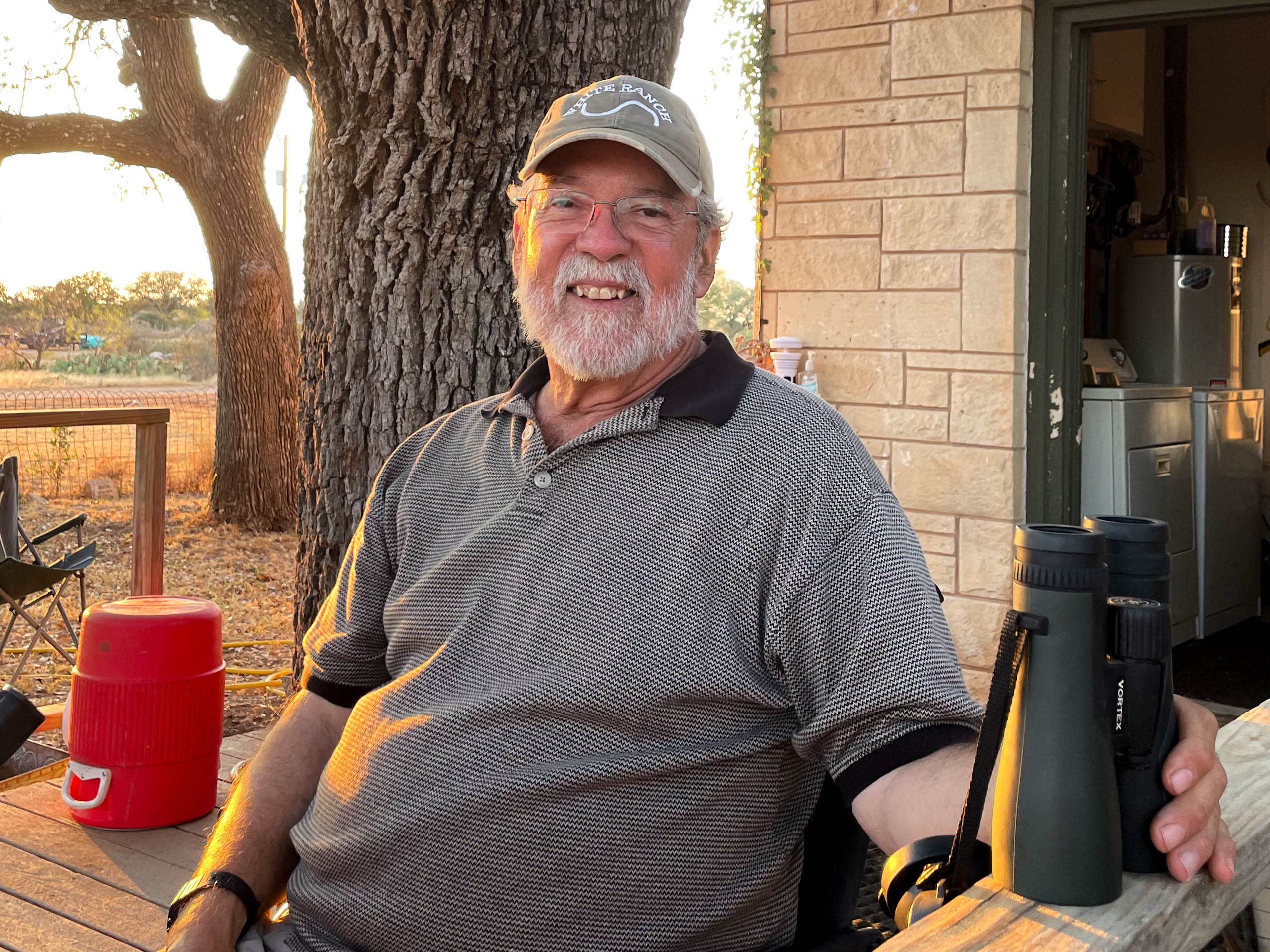 Portrait of Brian Wright smiling on his back porch wearing a White Ranch baseball cap and holding a pair of binoculars.