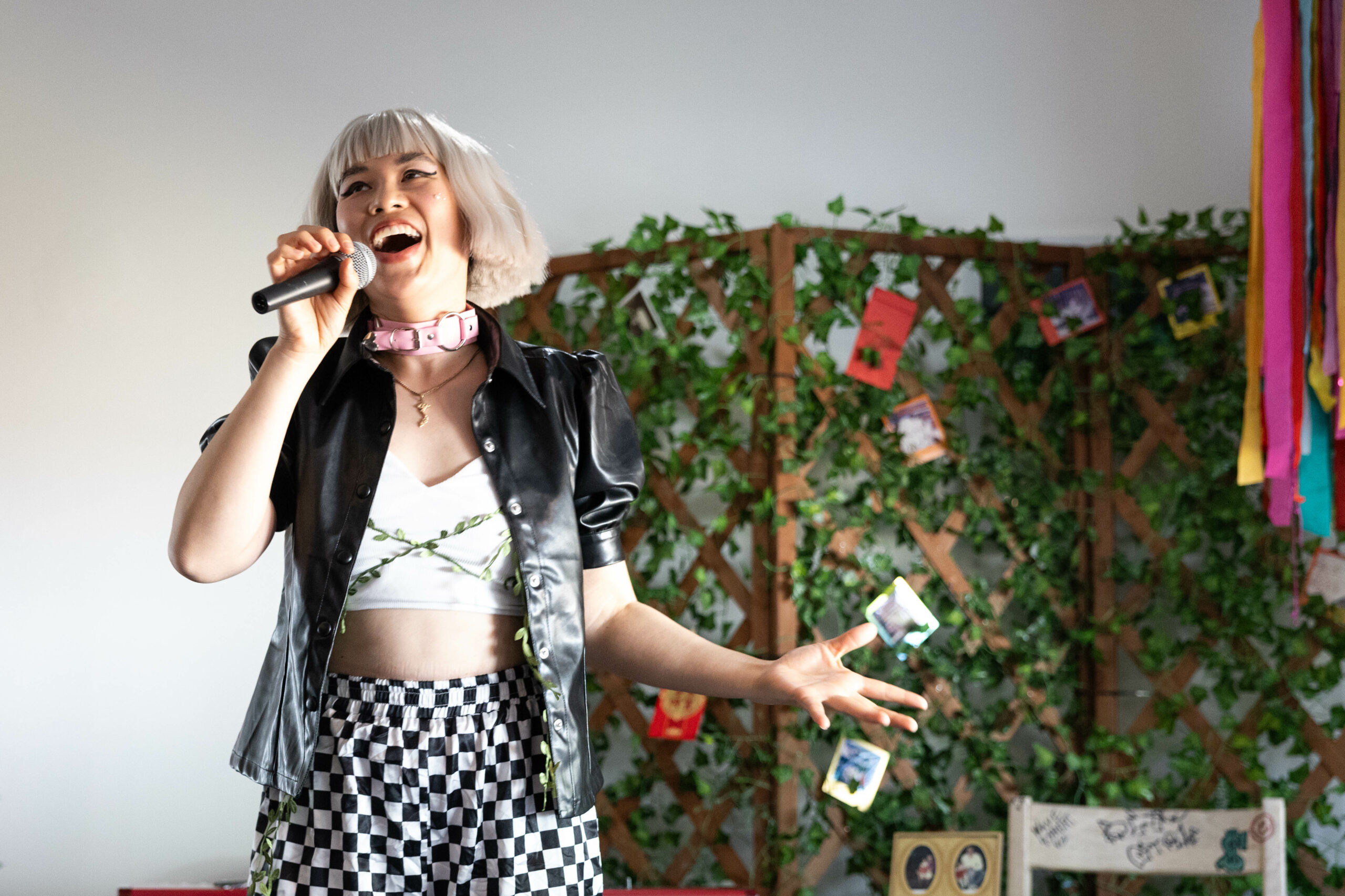 Promqueen smiles and sings into a microphone in front of a colorful backdrop. She wears a pink choker, black and white checkered pants and a black leather puff-sleeved jacket over a white top with green vine wrapped around her body.